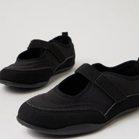 A pair of black Mary Jane-style slip-on shoes with hook-and-loop strap closures, photographed on a clean light gray studio background.