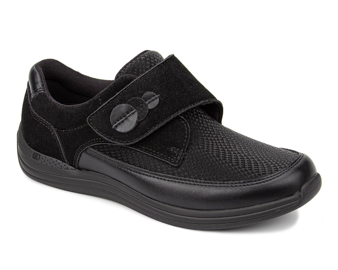 A black slip-on shoe with a textured mesh upper, a wide hook-and-loop strap with a circular button closure, and a thick black sole, shown on a white background.