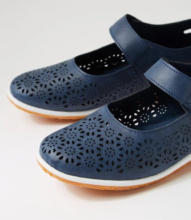A pair of navy blue Mary Jane-style shoes with floral laser-cut perforations on the toe and upper straps, featuring a white midsole and a natural brown outsole, shown on a light gray background.
