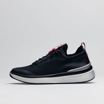 One of a pair of BALA Twelves in Nocturnal, which are premium, athletic-style sneakers for nurses and healthcare workers. The shoe has a black upper, white sole, and silver detailing around the heel, along with black laces and bright red and sky blue deta