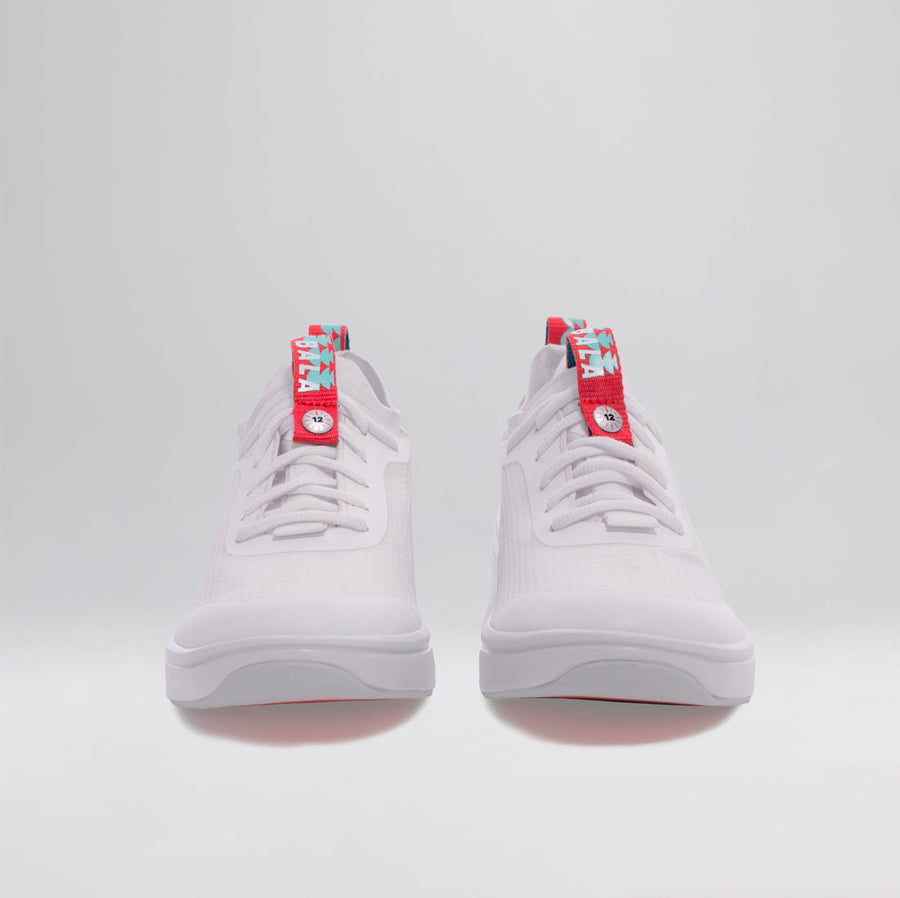 A pair of BALA Twelves in Flow White, which are bright white athletic-style sneakers for nurses and healthcare workers. They are photographed from the front; the tongue features a bright red and sky blue ribbon that snaps over white laces to keep them in place. The ribbon is fastened with a silver button, designed to look like a clock, with the numeral 12 in the center.