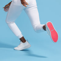 A Black woman runs in a pair of BALA Twelves in Flow White, which are premium, athletic-style sneakers for nurses and healthcare workers. The woman is only visible from the hips down. Her hands move alongside her hips, adorned with gold rings and bracelets, and long polished nails. She is wearing white, jogger-style scrub bottoms. One shoe is on the ground, the other is in the air, showcasing the bright red sole and its sky blue BALA logo. The photo is taken against a solid, sky-blue background.
