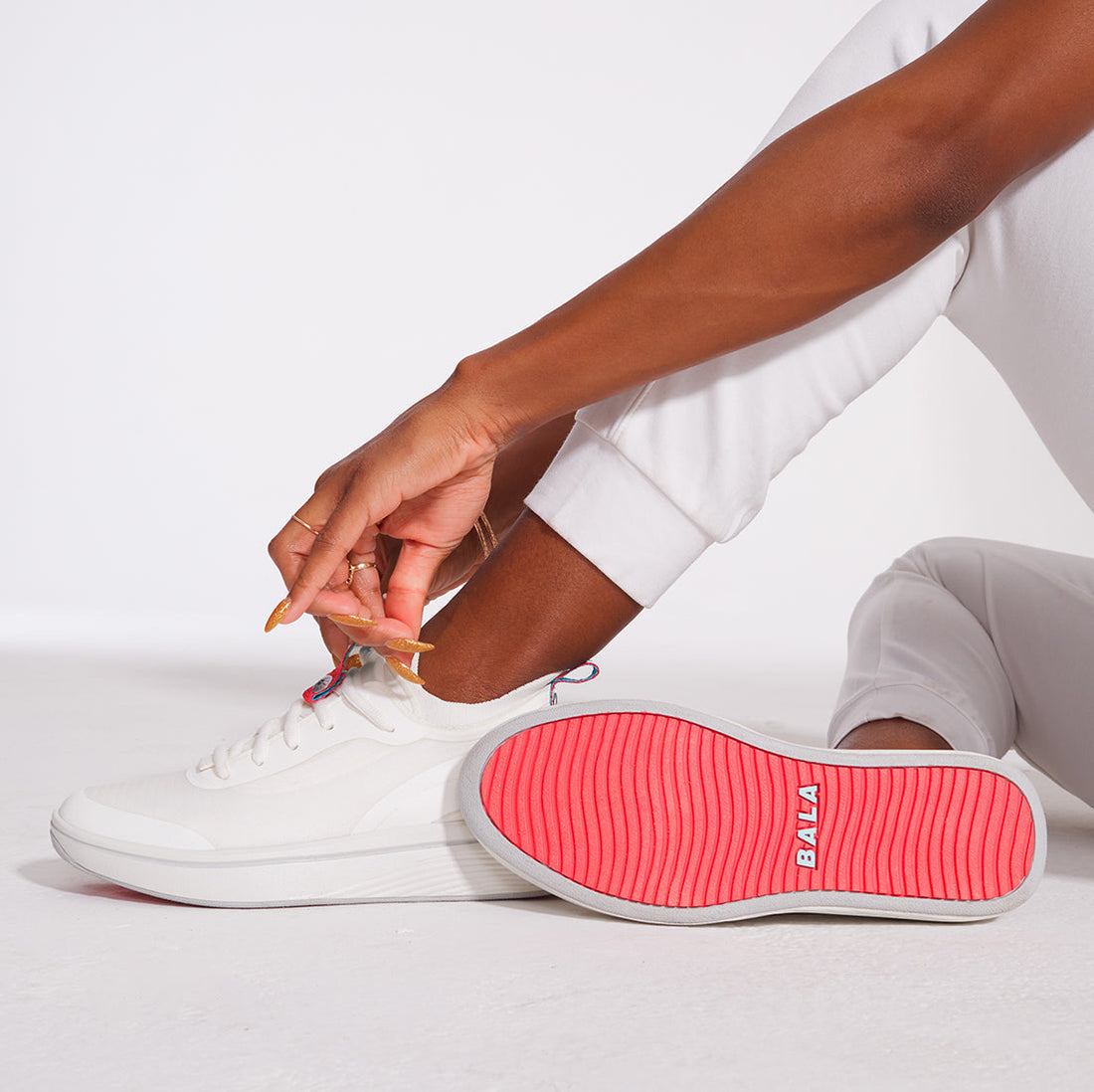 A Black woman sits on the floor to adjust her BALA Twelves in Flow White, which are bright white athletic-style sneakers for nurses and healthcare workers. Her hands are adorned with gold rings and bracelets, and long, polished nails. She is wearing white, jogger-style scrub bottoms. One foot is on the floor; she is adjusting the tongue of the shoe on this foot. Her other leg falls to the side so that the sole of her shoe is visible; it has a bright red sole with a sky blue BALA logo.